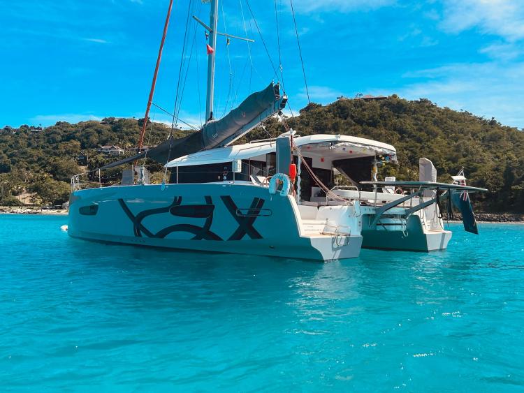 2024 Excess 14 catamaran anchored in clear turquoise waters, lush green hills in background.